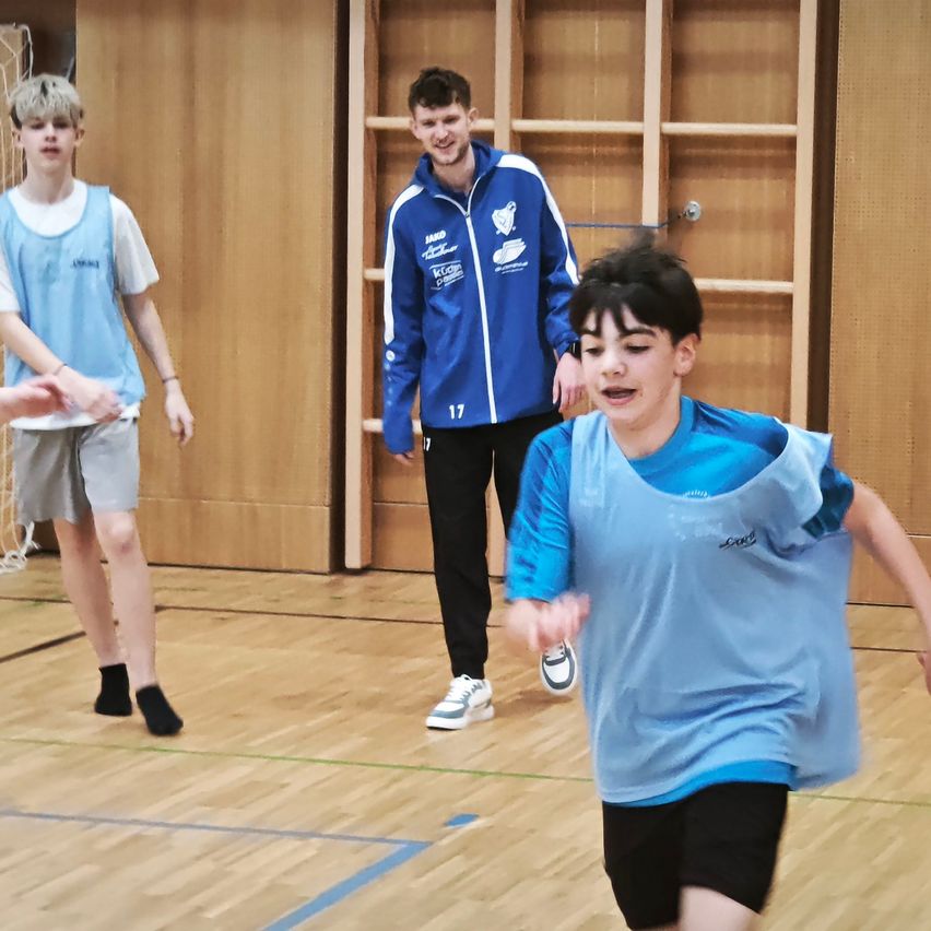 Drei Jungen in blauen Uniformen spielen ein Spiel drinnen. Ein Junge läuft vorwärts, ein anderer schaut zu und ein dritter steht hinter ihnen in der Nähe einer Holzwand.