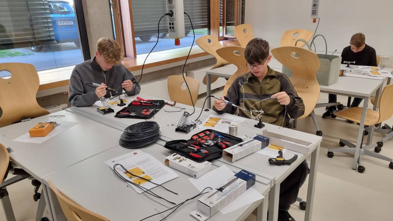 Zwei Jungen arbeiten an einem Lötprojekt an einem Tisch mit verschiedenen Werkzeugen und Drähten. Ein Junge benutzt ein Löteisen, während der andere sich auf eine Leiterplatte konzentriert.
