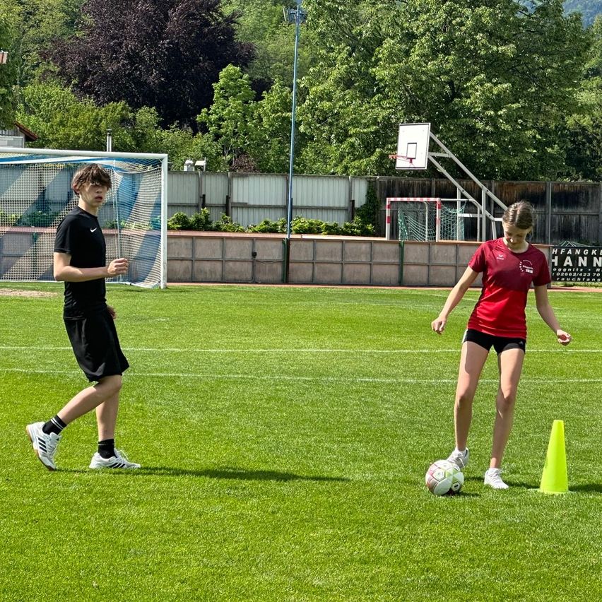 Zwei Personen spielen Fußball auf einem Feld, ein junger Mann in Schwarz und eine junge Frau in Rot. Sie stehen in der Nähe eines gelben Kegels, was auf ein Übungsspiel hinweist. Im Hintergrund befinden sich ein Fußballtor und ein Basketballkorb, was auf einen Mehrzweck-Sportbereich hindeutet.