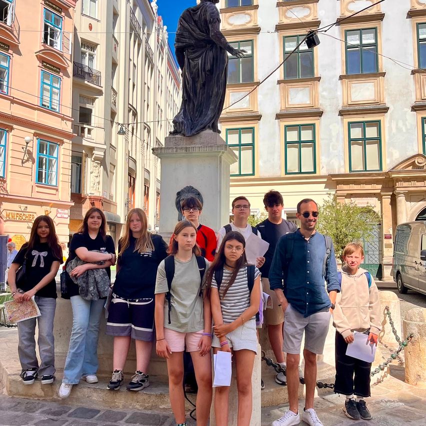 Eine Gruppe von Menschen steht vor einer Statue in einer städtischen Umgebung mit mehreren Gebäuden im Hintergrund. Sie tragen Freizeitkleidung und scheinen für ein Foto zu posieren.
