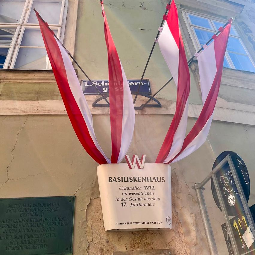 Fahnen und ein Schild für Basiliskenhaus, ein historisches Gebäude, sind an einer Wand angebracht. Das Gebäude wurde 1212 gegründet. Es befindet sich in der 1. Schottengasse.