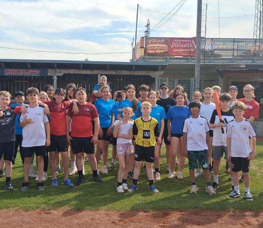 Eine Gruppe von Kindern und Teenagern posiert für ein Foto auf einem Baseballfeld, einige tragen Trikots und halten Baseballschläger. Im Hintergrund ist ein Banner mit dem Text 'Wilczek' zu sehen.