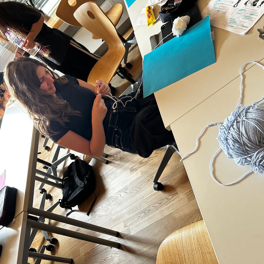 Two women are indoors. One woman is seated on the floor and knitting while the other stands behind her. They are surrounded by tables, chairs, and a backpack.