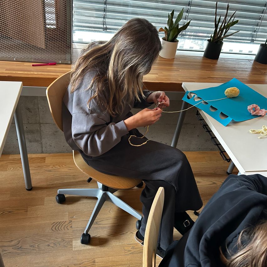 A woman sits at a wooden chair, focused on knitting with yarn on a blue mat. She is seated in an office with a desk and plants in the background.
