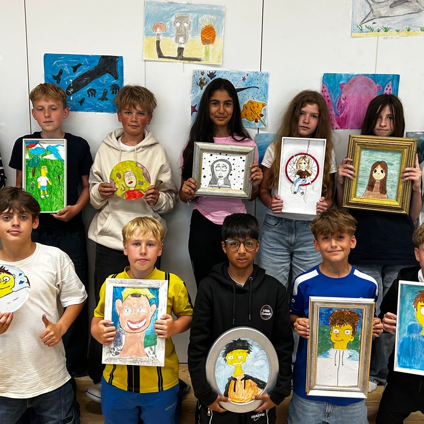 Eine Gruppe von Kindern hält gerahmte Zeichnungen von Gesichtern und posiert für ein Foto in einem Klassenzimmer.