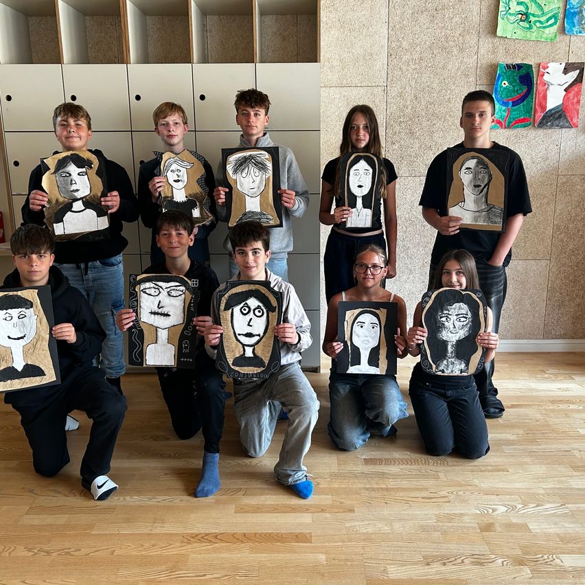 Eine Gruppe von Schülern posiert mit ihren Porträtzeichnungen in einem Klassenzimmer, alle lässig gekleidet.