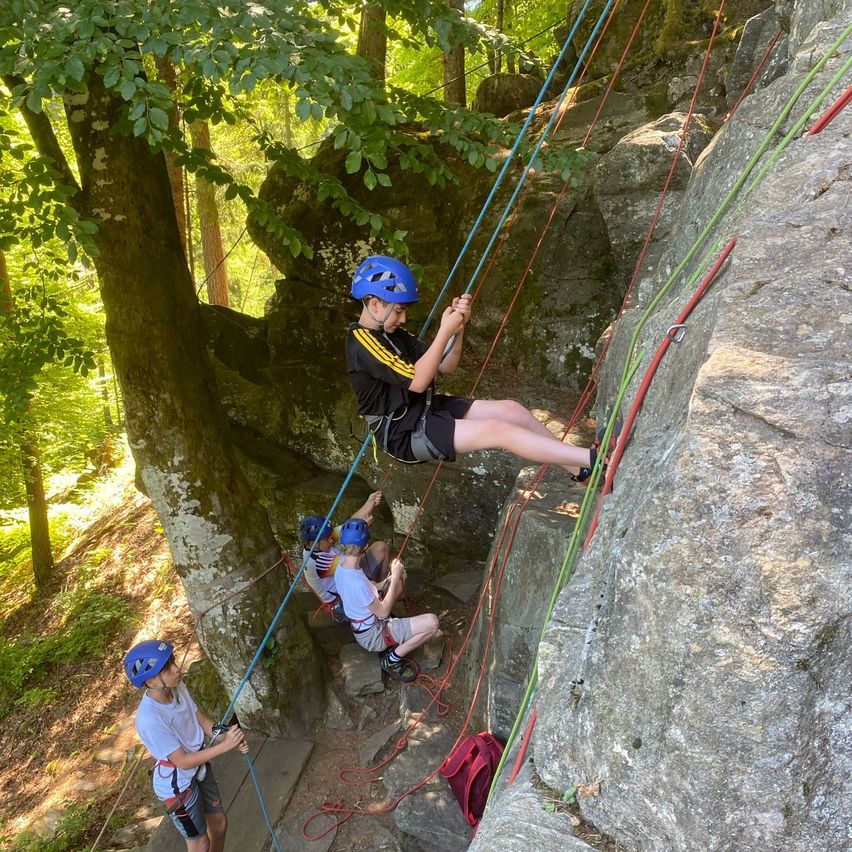 Vier Kinder klettern mit Seilen und Helmen an einem Felsen, ein Kind mit blauem Helm ist halb den Felsen hoch.