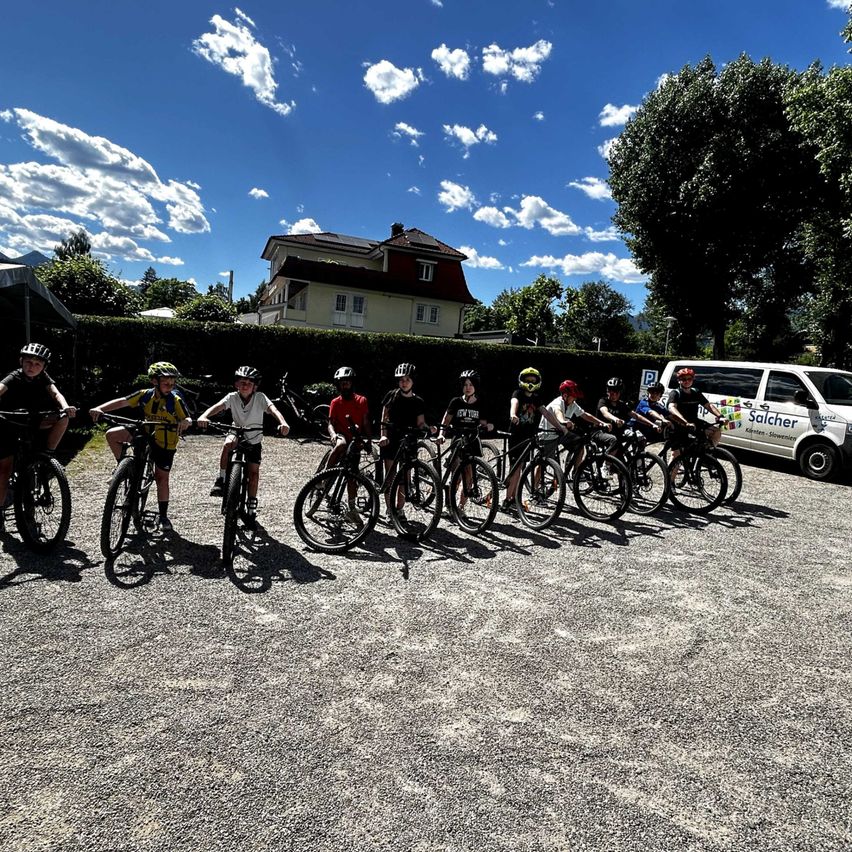 Eine Gruppe von Radfahrern mit Helmen steht in einer Reihe, mit einem weißen Van, der dahinter geparkt ist. Die Radfahrer tragen verschiedenfarbige Trikots, und im Hintergrund befinden sich ein Haus und Bäume.