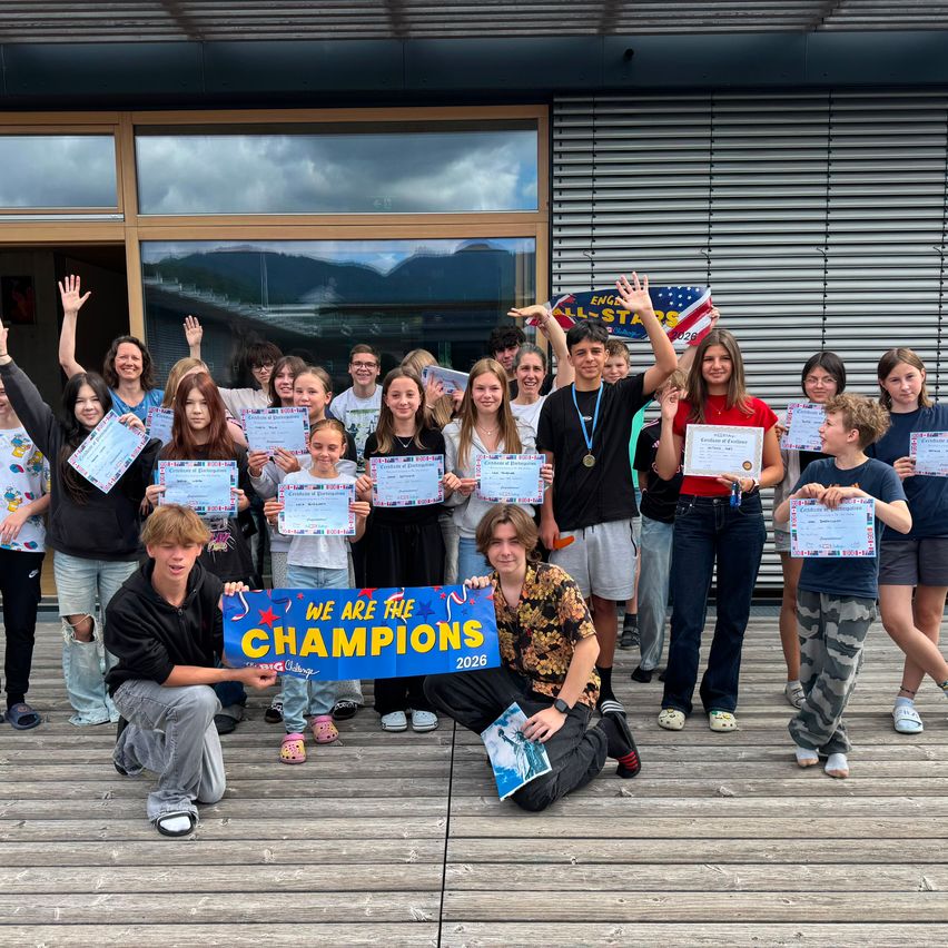 Eine Gruppe von Schülern posiert für ein Foto, hält Urkunden und ein Schild mit der Aufschrift 'Wir sind die Champions 2026'. Sie stehen vor einem Gebäude mit einer Metallfassade.