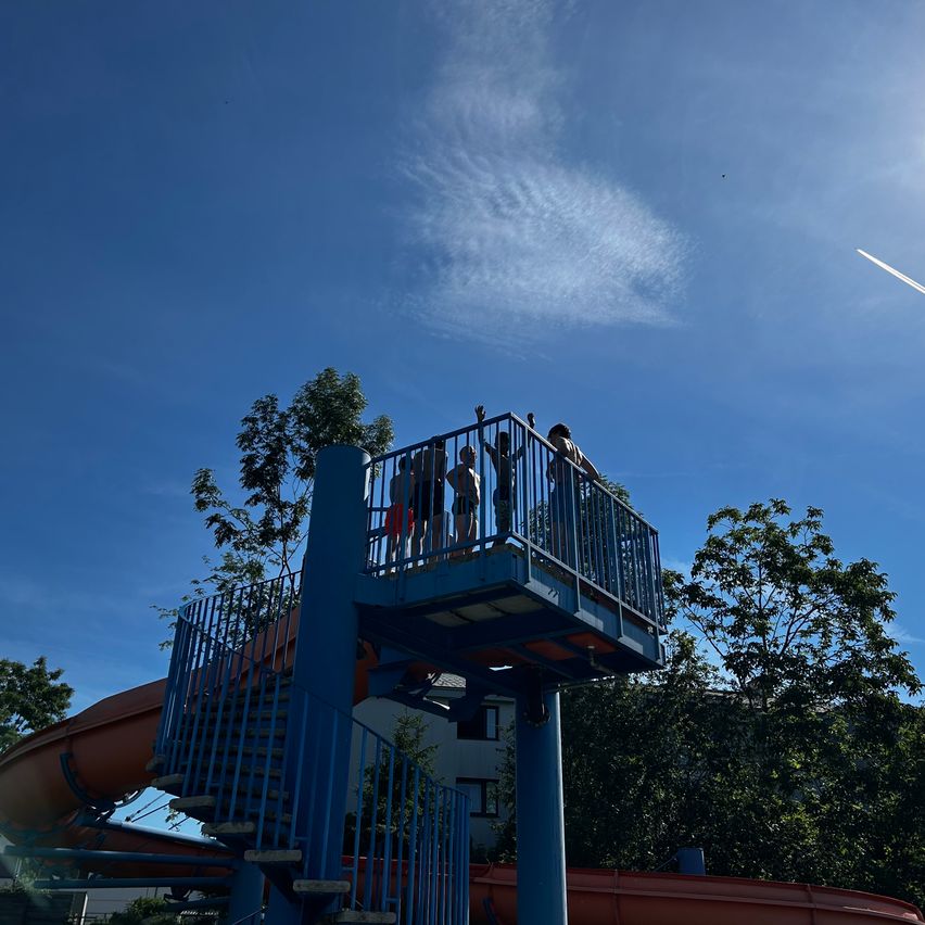 Eine Gruppe von Menschen steht auf einer Aussichtsplattform über einer Wasserrutsche. Bäume und ein blauer Himmel sind im Hintergrund.