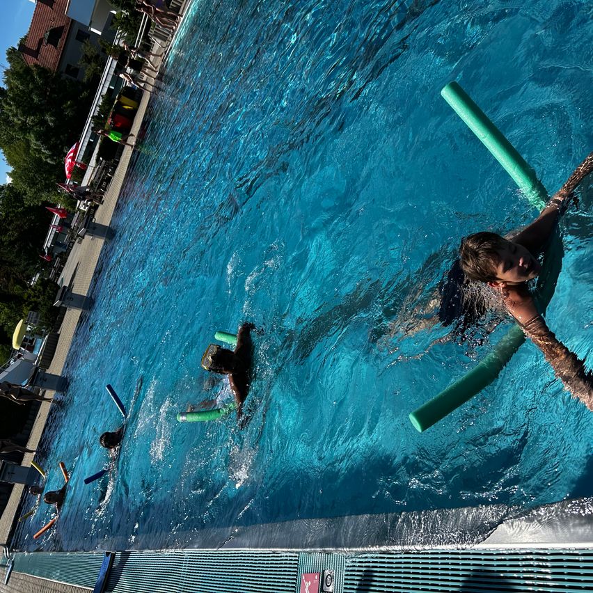 Kinder schwimmen in einem großen Außenpool. Einer hält einen grünen Schwimmreifen, andere schwimmen mit Schwimmbrillen. Bäume und ein Gebäude sind im Hintergrund zu sehen.