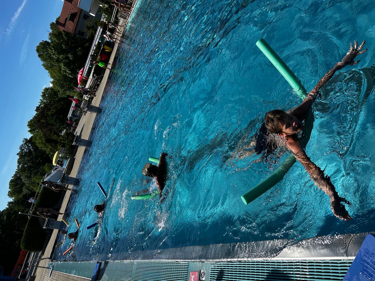 Kinder schwimmen in einem großen Außenpool. Einer hält einen grünen Schwimmreifen, andere schwimmen mit Schwimmbrillen. Bäume und ein Gebäude sind im Hintergrund zu sehen.