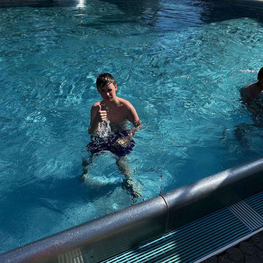Ein junger Junge zeigt im Schwimmbecken einen Daumen nach oben, eine weitere Person ist im Hintergrund.