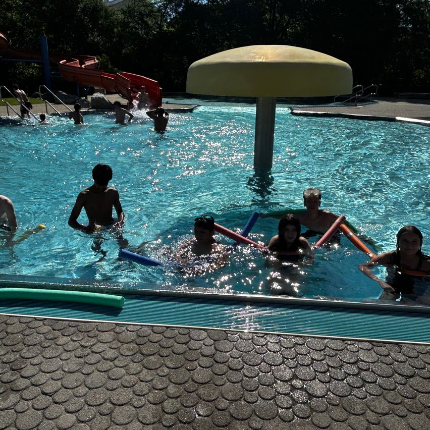 Eine Gruppe von Menschen schwimmt in einem Pool mit einer Rutsche im Hintergrund. Einige benutzen Schwimmhilfen.