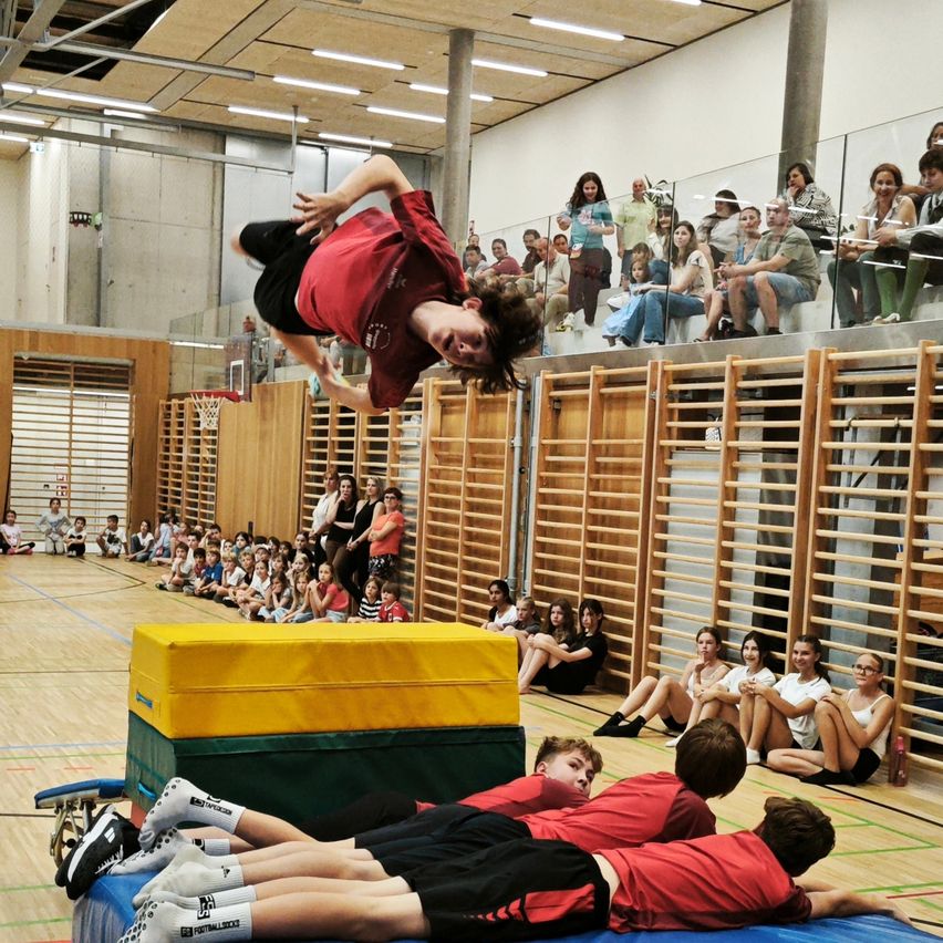 Ein junger Junge führt einen akrobatischen Überschlag über einem Gymnastiktisch durch, während eine Menge von den Tribünen zuschaut.