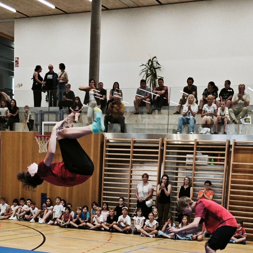 Ein Indoor-Basketballplatz, eine Person führt einen Trick vor, während mehrere Personen zuschauen. Eine Gruppe von Kindern sitzt auf dem Boden. Ein Mann in Rot versucht, den Performer zu fangen.