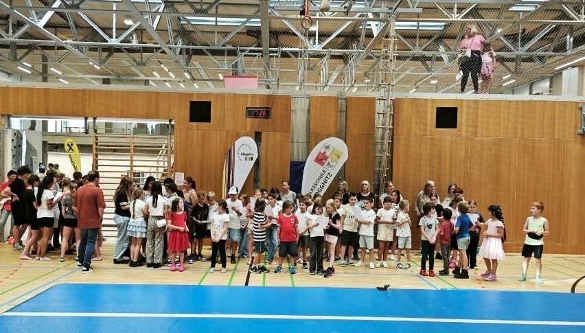 Viele Kinder und Erwachsene sind in einer Sporthalle mit blauen Böden versammelt. Einige stehen, andere sitzen auf den Tribünen. Die Wände sind mit Bannern dekoriert.