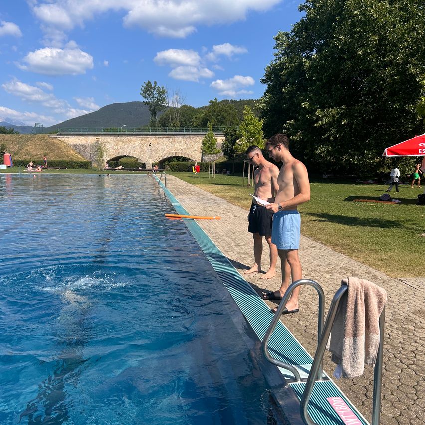 Zwei Männer in Badekleidung stehen an einem Schwimmbecken. Ein Schwimmer taucht ins Wasser. Dahinter sind eine Brücke und eine Bergkette zu sehen.
