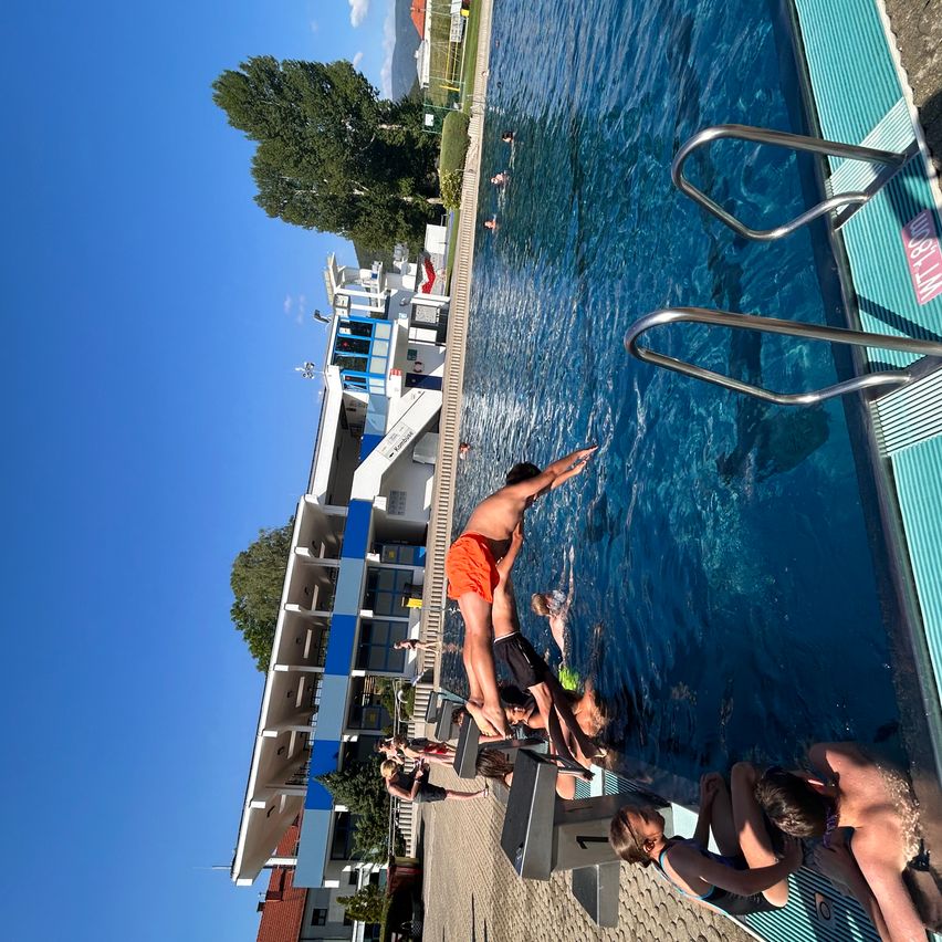 Luftaufnahme eines Schwimmbeckens mit Schwimmern und anderen Personen am Rand. Das Becken hat blaues Wasser und Metallhandläufe. In der Nähe befinden sich Gebäude und ein Baum ist im Hintergrund sichtbar.