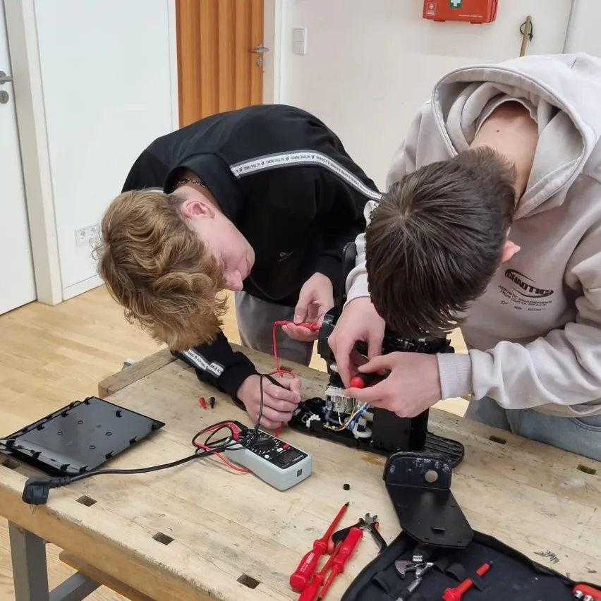 Zwei junge Männer arbeiten an einem elektronischen Projekt auf einem Holztisch. Einer beugt sich vor und der andere steht, beide konzentrieren sich auf die Aufgabe.