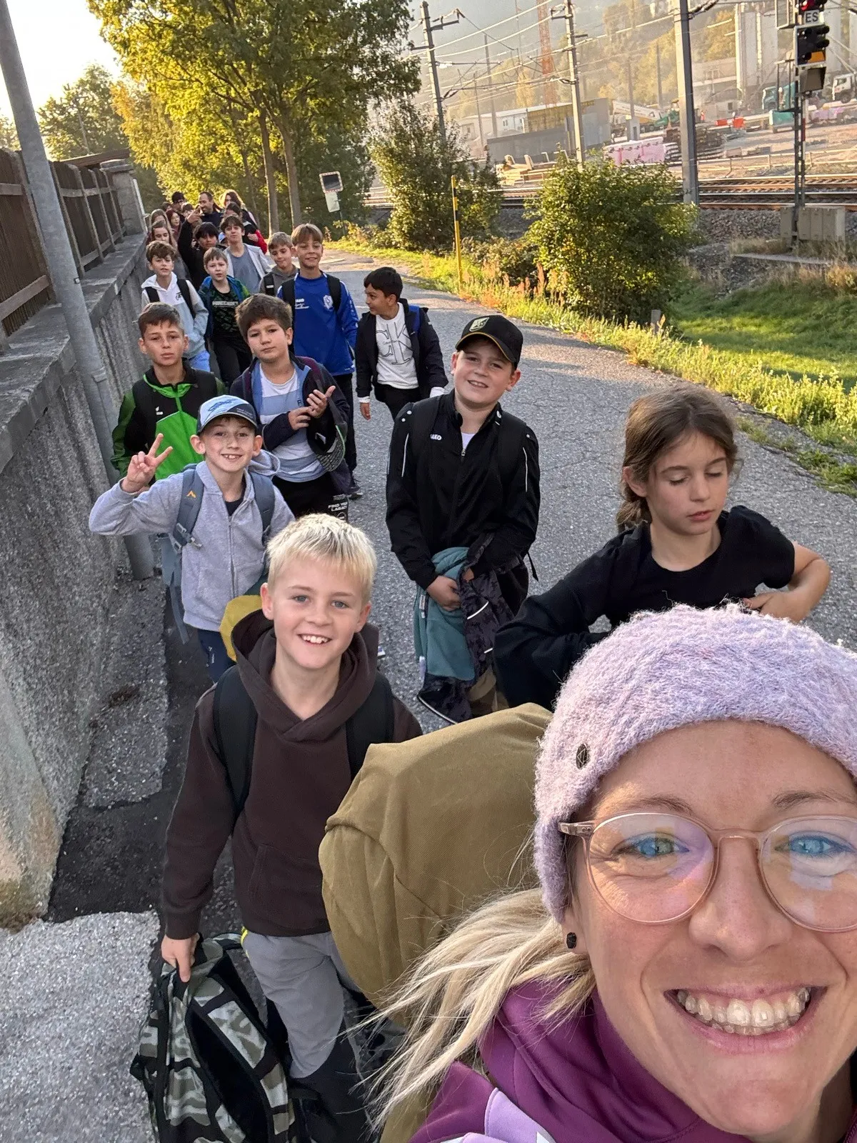 Eine Gruppe von Kindern und einer Frau geht auf einem Bürgersteig. Die Kinder tragen Rucksäcke und einige tragen Mützen. Die Frau trägt eine Brille und eine Mütze.