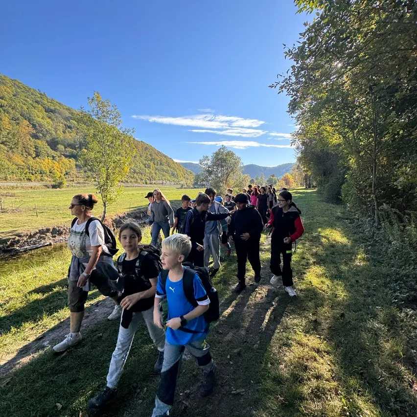 Eine Gruppe von Menschen, einschließlich Kindern, geht auf einem Grasweg in einem ländlichen Gebiet. Sie tragen Rucksäcke und einige tragen Hüte. Der Weg ist von Bäumen und Gras umgeben, mit einem Fluss und Bergen im Hintergrund.