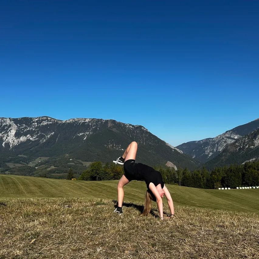 Eine Frau macht einen Handstand auf einer Wiese mit Bergen im Hintergrund.
