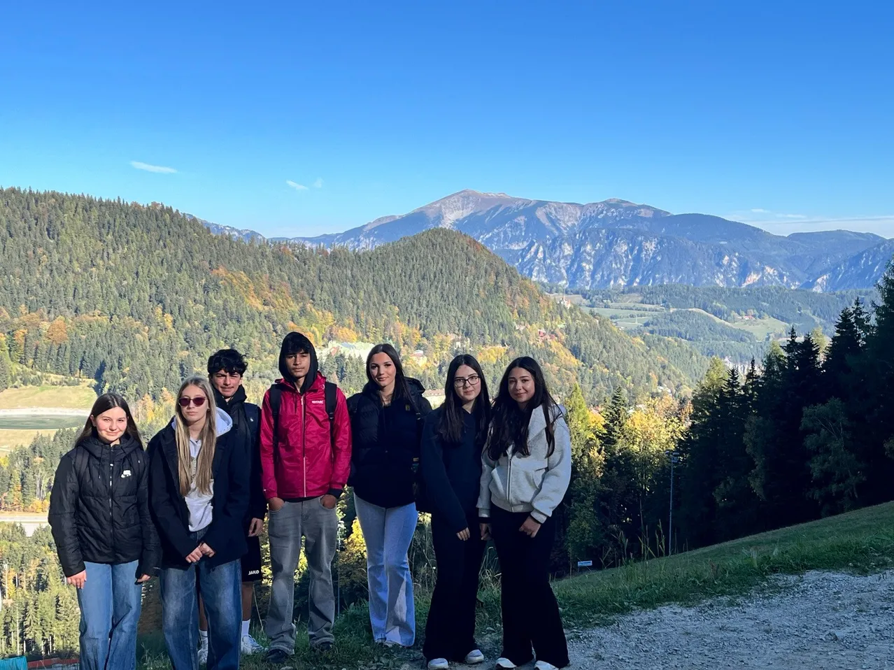 Eine Gruppe junger Menschen steht auf einem Bergpfad und lächelt und posiert für ein Foto. Der Hintergrund zeigt eine wunderschöne Bergkette mit Bäumen und einem klaren blauen Himmel.