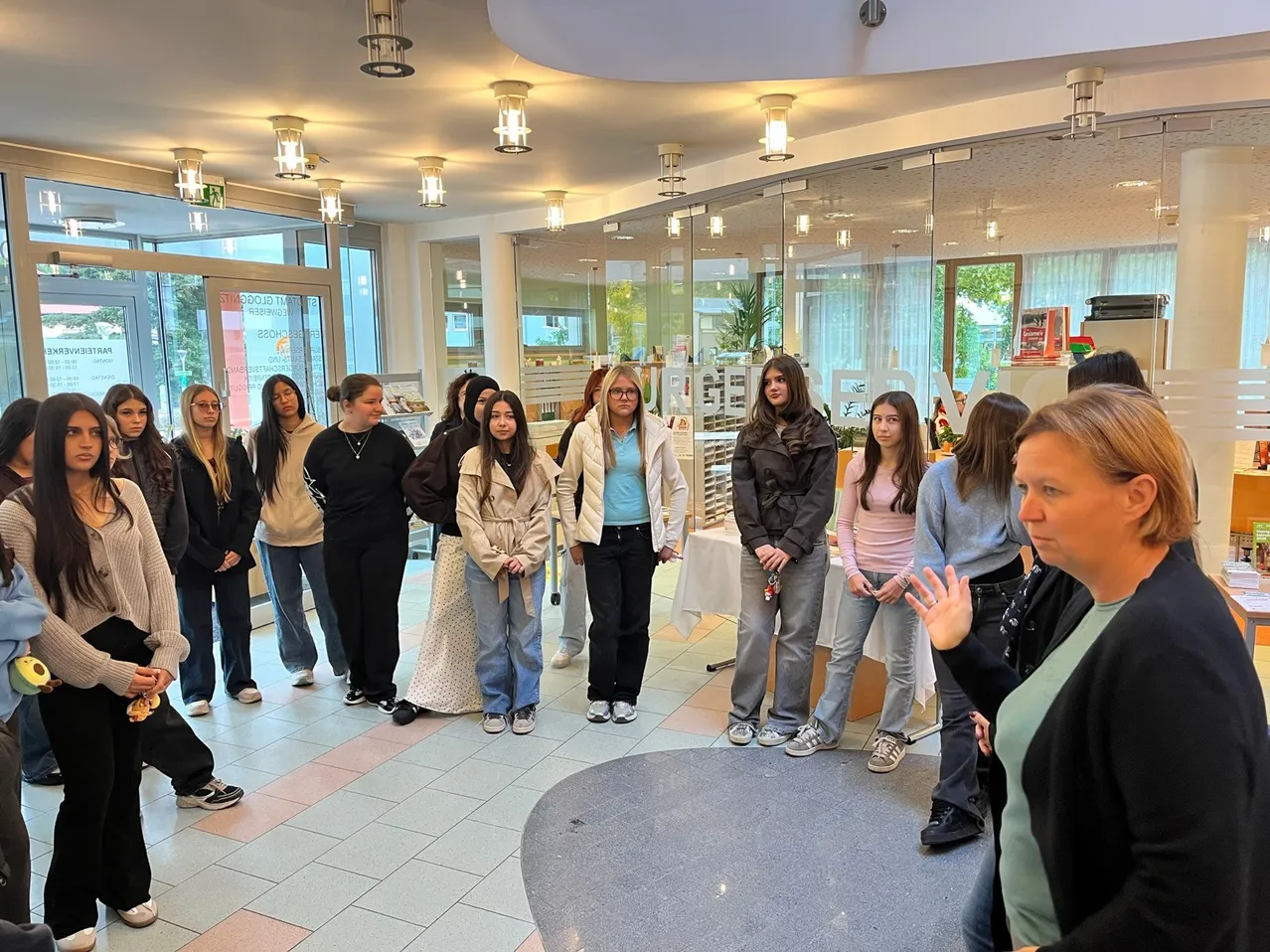 Eine Gruppe junger Frauen und eine Frau in einem schwarzen Blazer stehen in einem Raum mit Glaswänden und Deckenleuchten.