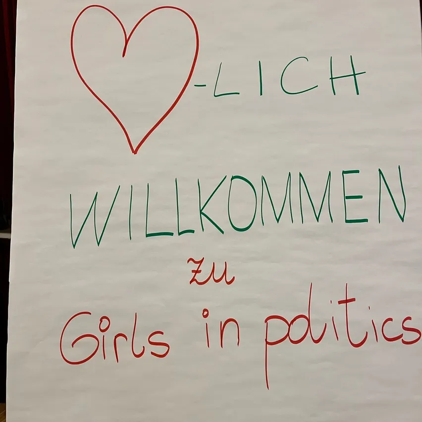 Ein weißes Brett mit einem roten Herzen und grünem Text, der 'LICH WILLKOMMEN zu Girls in politics' lautet.