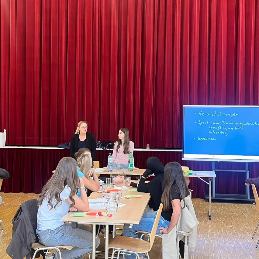 Eine Gruppe von Frauen sitzt um einen Tisch in einem Klassenzimmer. Eine Frau steht vorne, in der Nähe einer Projektionsleinwand mit Text.