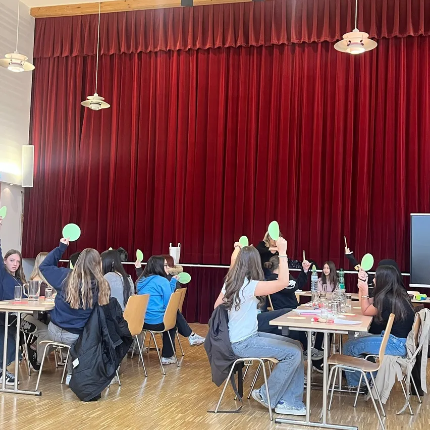 Eine Gruppe von Menschen sitzt an Tischen in einem Raum mit Holzböden und einem roten Vorhang im Hintergrund. Sie halten grüne Karten und scheinen an einer Aktivität beteiligt zu sein.