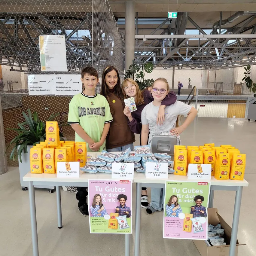 Vier Kinder stehen hinter einem Tisch mit gelben Snacks. Der Tisch hat zwei Poster mit dem Text 'Tu Gutes für dich & mich'.