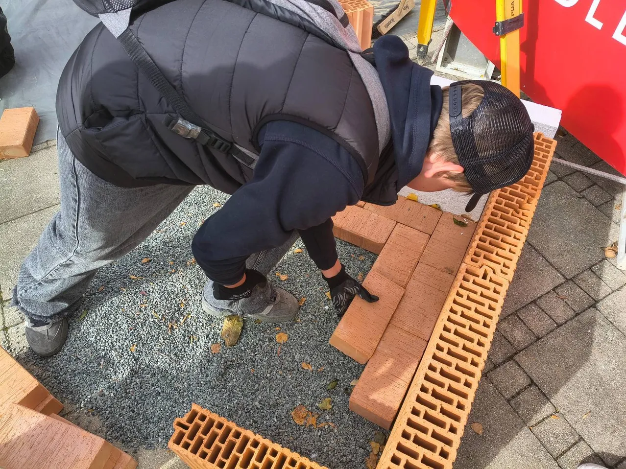 Eine Person, die eine schwarze Jacke, graue Hose und eine schwarze Kappe trägt, beugt sich vor und legt einen Ziegelstein auf eine Ziegelmauer.