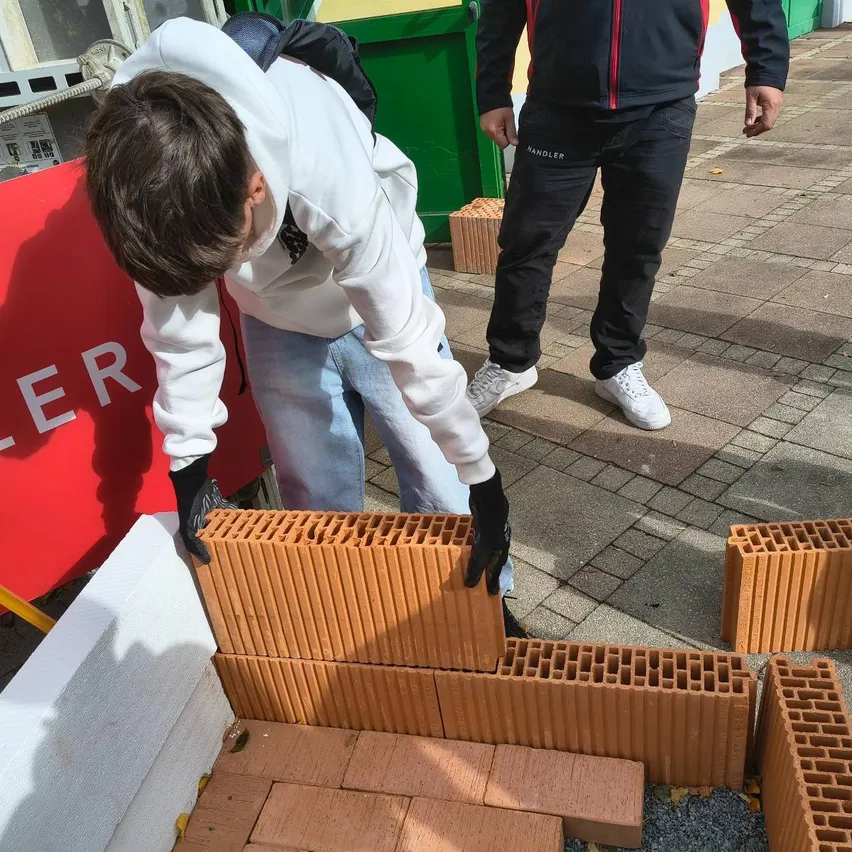 Ein Mann arbeitet an einer Ziegelmauer, trägt eine weiße Kapuzenjacke, Jeans und schwarze Handschuhe. Er beugt sich vor und legt Ziegelsteine auf die Mauer. Ein anderer Mann steht hinter ihm, trägt eine schwarze Jacke, Hose und weiße Turnschuhe.
