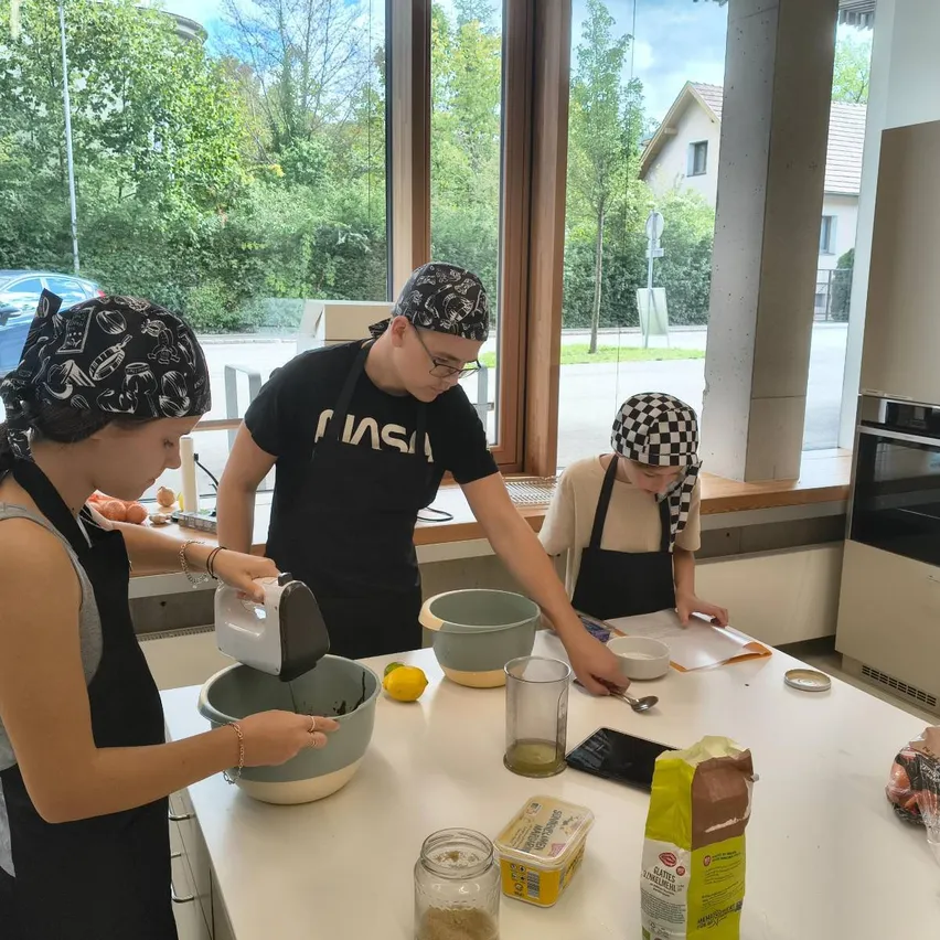Drei Personen bereiten in einer Küche Essen zu. Eine Frau gießt Zutaten in eine Schüssel, ein Mann steht hinter ihr und eine andere Frau sitzt am Tisch.