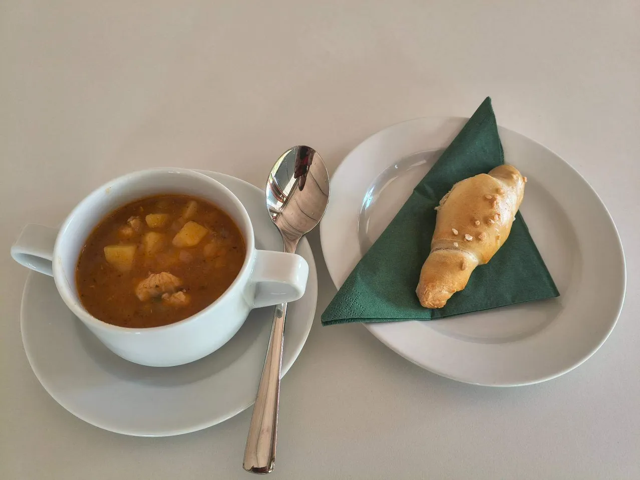 Eine weiße Tasse Suppe mit einem Löffel auf einer Untertasse, neben einem Teller mit einem Croissant auf einem grünen Serviettentuch.