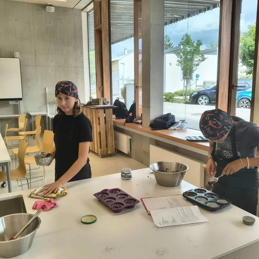Drei junge Mädchen sind in einer Küche, tragen Stirnbänder und Schürzen. Sie bereiten Essen zu, wobei ein Mädchen Geschirr spült, ein anderes Zutaten mischt und das dritte Zutaten in eine Schüssel gibt. Es gibt verschiedene Küchenutensilien und Zutaten auf dem Tisch, und eine Tafel ist an der Wand montiert.