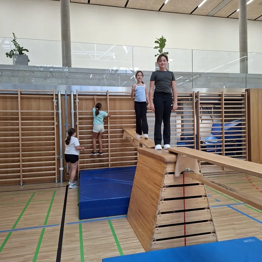 Drei Mädchen üben das Gleichgewicht auf einem Holzbalken in einer Turnhalle. Ein Mädchen steht auf dem Balken, während die anderen beiden auf dem Boden stehen.