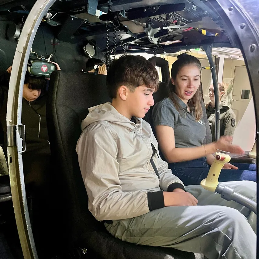 Ein Junge sitzt in einem Hubschraubersimulator, trägt ein Headset und hält einen Steuerknüppel. Eine Frau sitzt neben ihm und hält ebenfalls einen Steuerknüppel.
