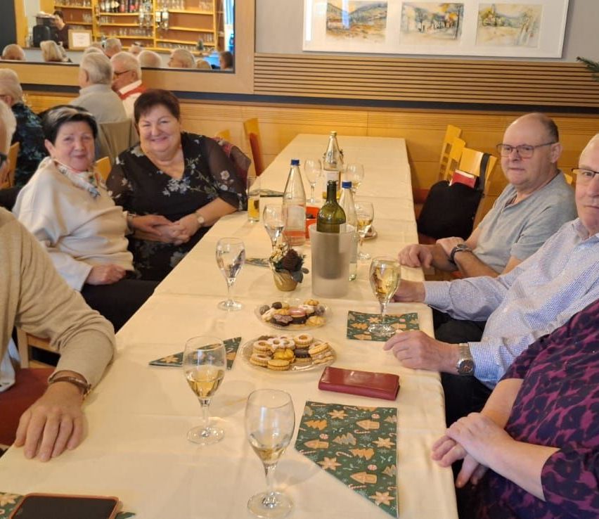 Mehrere ältere Erwachsene sitzen um einen Tisch herum, lächeln und unterhalten sich. Der Tisch ist mit Weingläsern, Tellern mit Desserts und Weinflaschen gedeckt. Hinter ihnen zeigt eine Wand gerahmte Bilder.