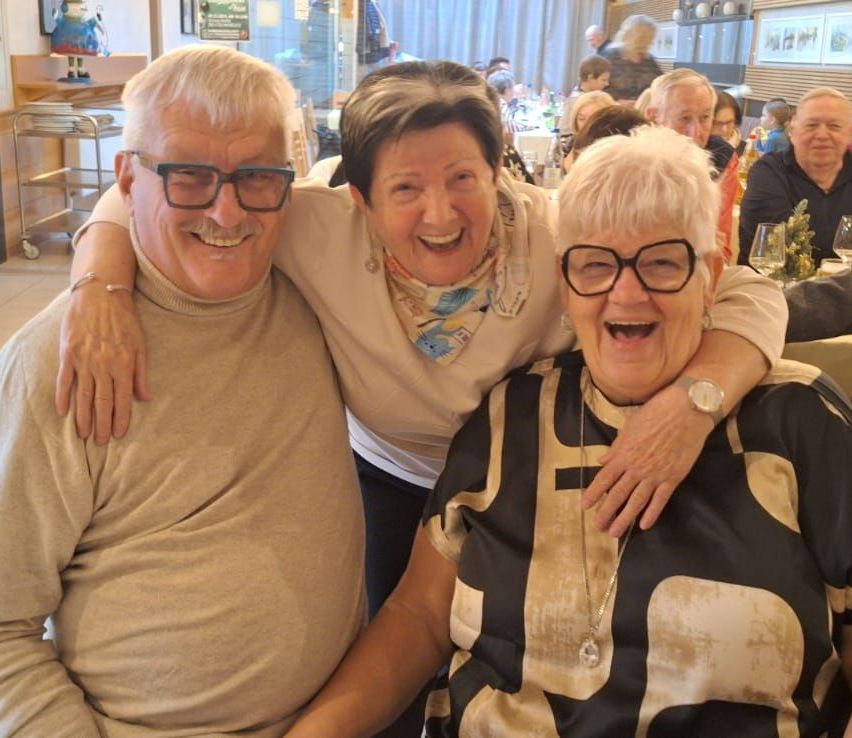Drei ältere Personen, zwei Frauen und ein Mann, lächeln und posieren für ein Foto. Der Mann hat seinen Arm um die Frau auf seiner rechten Seite gelegt. Dahinter sitzen mehrere Personen an Tischen.