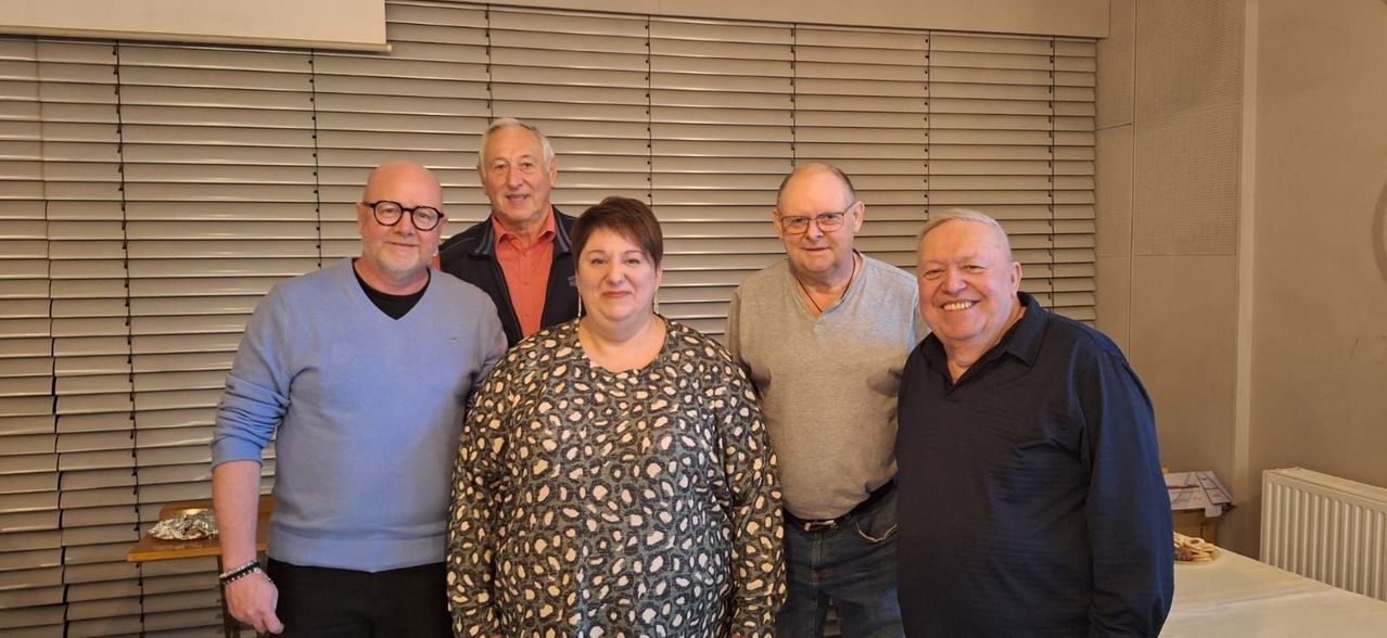 Fünf Menschen posieren für ein Foto. Sie stehen vor einem Fenster mit Jalousien. Die Frau in der Mitte trägt ein Leopardenmuster-Hemd. Der Mann rechts trägt ein schwarzes Polohemd. Der Mann links trägt einen blauen Pullover. Der Mann in der Mitte trägt eine Jacke und ein orangefarbenes Hemd.