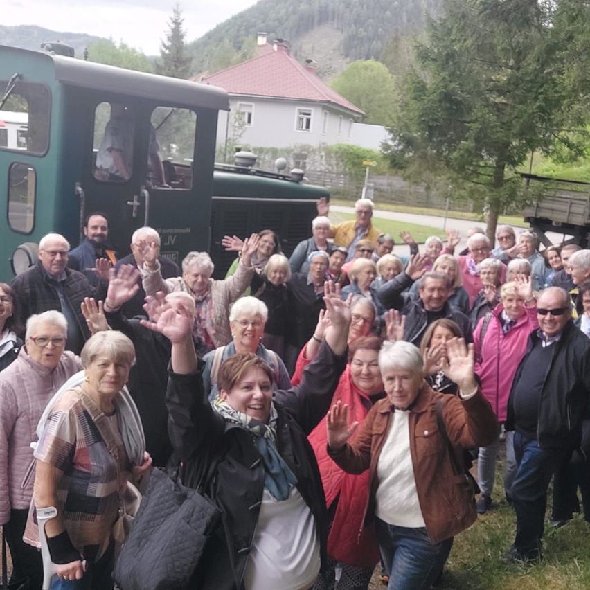 Eine Gruppe älterer Menschen steht vor einem grünen Zug und winkt. Ein Haus mit rotem Dach befindet sich hinter ihnen.