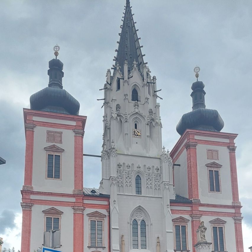 Die Fassade einer großen Kirche mit zwei Türmen und einem zentralen Turm. Das Gebäude hat eine Mischung aus weißen und rosa Farben mit aufwendigen Verzierungen. Unter dem Fenster befindet sich eine Statue.