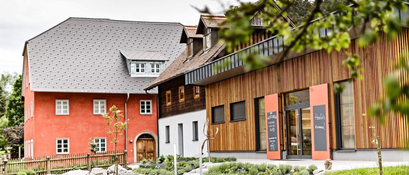 Zwei Gebäude mit modernem Design, eines rot mit einer Holztür und das andere mit holzfarbenem und schwarzem Äußeren, stehen auf einer grauen Kiesauffahrt. Sie haben Glastüren und Fenster.