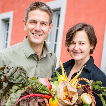 Ein Mann und eine Frau stehen lächelnd vor einem roten Gebäude und halten einen Korb voller frischem Gemüse und Obst.