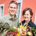 Ein Mann und eine Frau stehen lächelnd vor einem roten Gebäude und halten einen Korb voller frischem Gemüse und Obst.