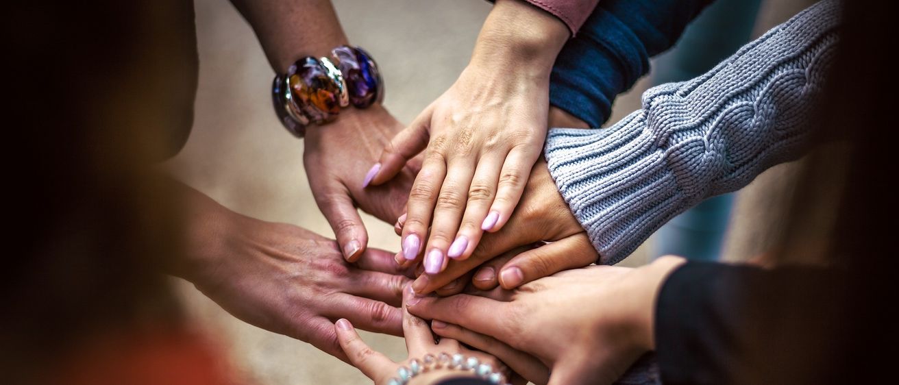 Eine Gruppe von Frauen legt die Hände ineinander, was Einheit und Teamarbeit zeigt. Ihre Hände sind mit rosa Nägeln bemalt.