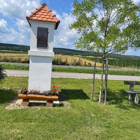 Eine kleine weiße Struktur mit einem roten Dach steht in einem Grasfeld. Es hat ein Fenster mit einem Metallgitter und einen Blumenkasten mit Blumen davor. Ein Baum und eine Bank sind in der Nähe.